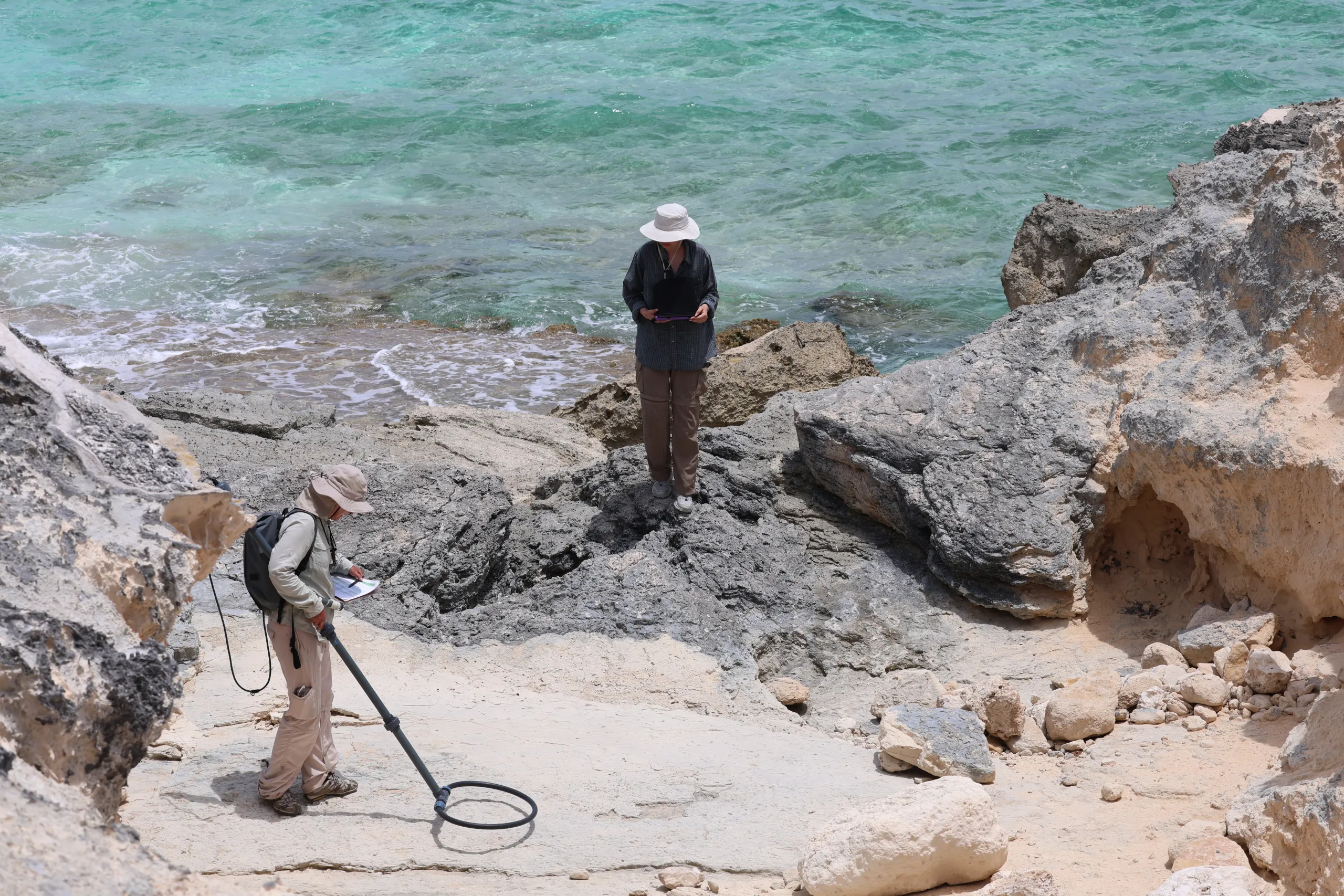 Two researchers wearing sun hats work along a rocky, light-colored shoreline beside bright turquoise ocean water. In the foreground, one person bends over holding a metal detector coil to the ground while reading papers. Slightly behind them on an elevated rock ledge, the second person stands holding a tablet or notebook.