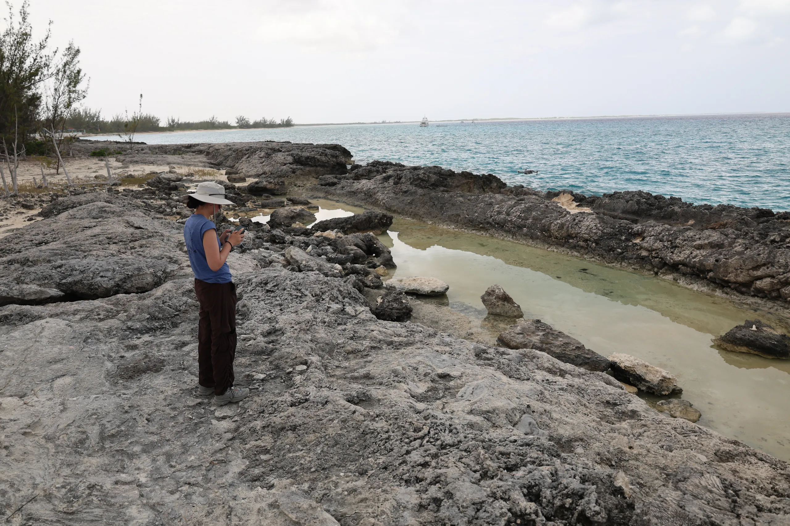 A person wearing a blue shirt, dark trousers, and a white sun hat stands on a dark, craggy limestone shoreline looking down at a mobile device or remote controller. Next to them is a tidal pool filled with shallow, still water, with turquoise ocean waters, distant boats, and a cloudy sky in the background.