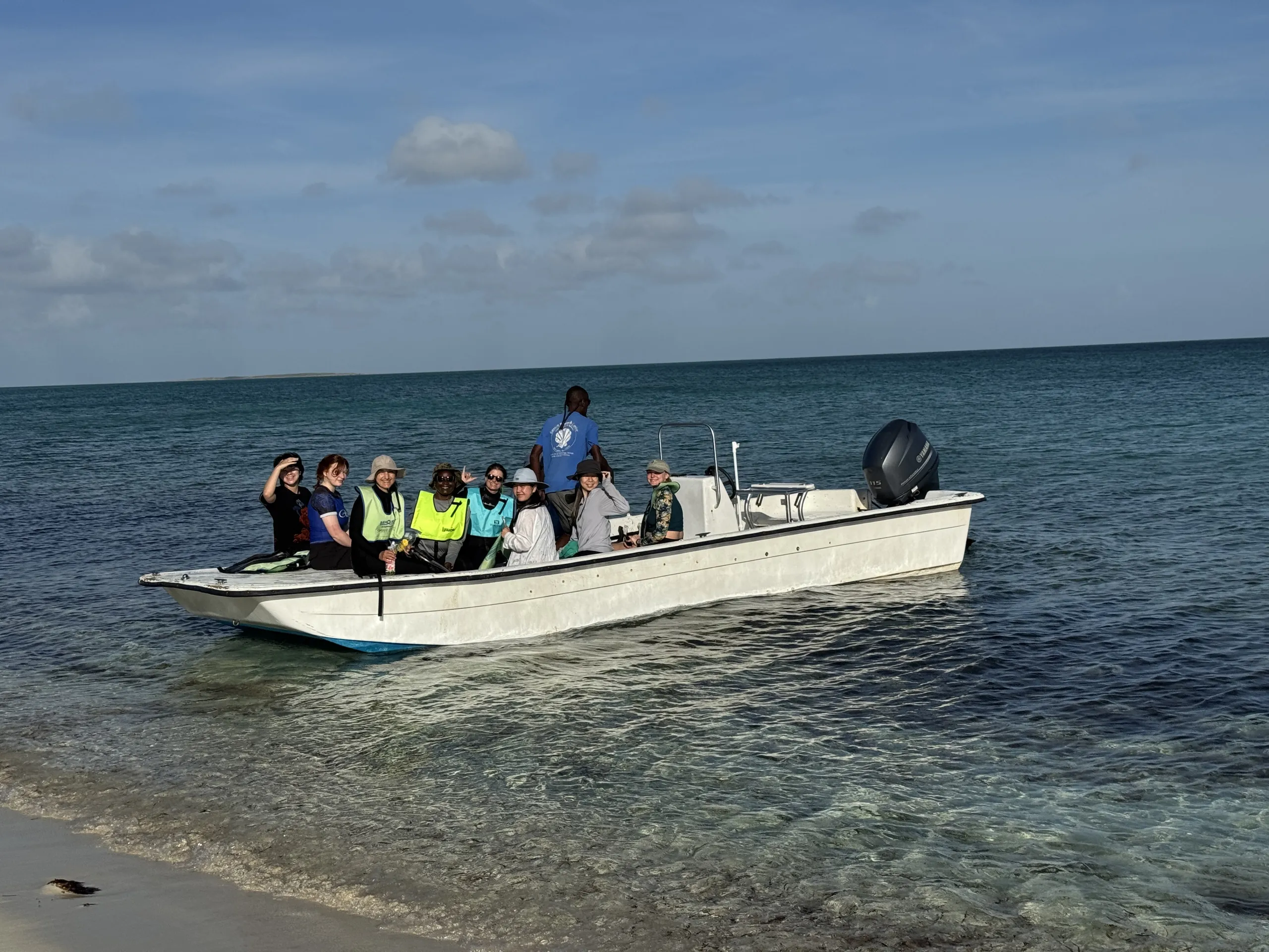 A group of eight people sit aboard a small white motorboat anchored in shallow, clear turquoise water near a sandy shoreline. Several passengers wear life jackets or sun hats, and one person stands near the stern steering position. In the background, calm open ocean extends to the horizon under a clear blue sky with light clouds.