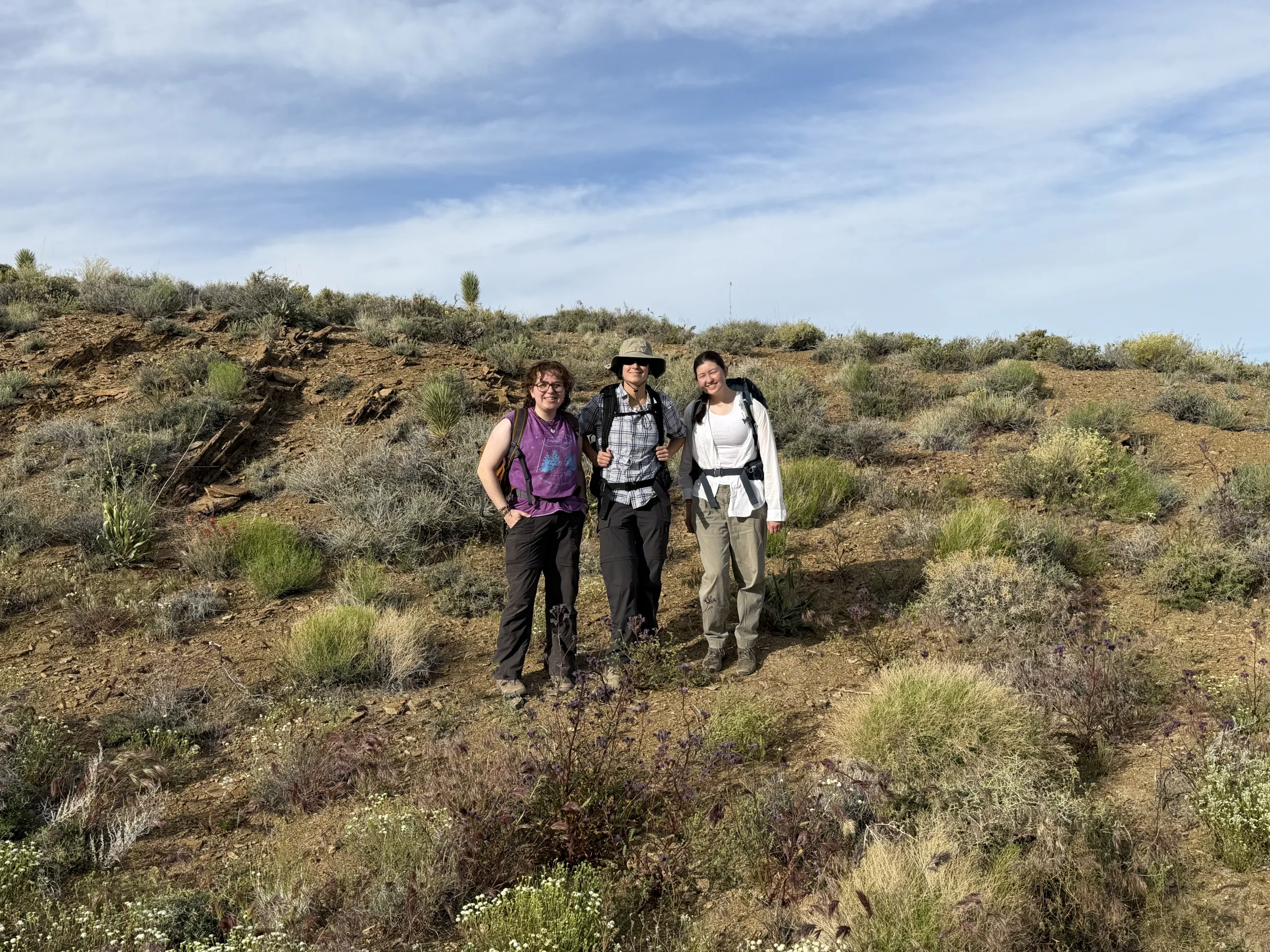 Three hikers wearing backpacks stand side by side on a dry, brush-covered hillside under a bright blue sky with light, wispy clouds. The person on the left wears a purple t-shirt, the person in the middle wears a plaid shirt and a wide-brimmed sun hat, and the person on the right wears a white top and cargo pants. Low desert shrubs and sparse vegetation cover the surrounding landscape.