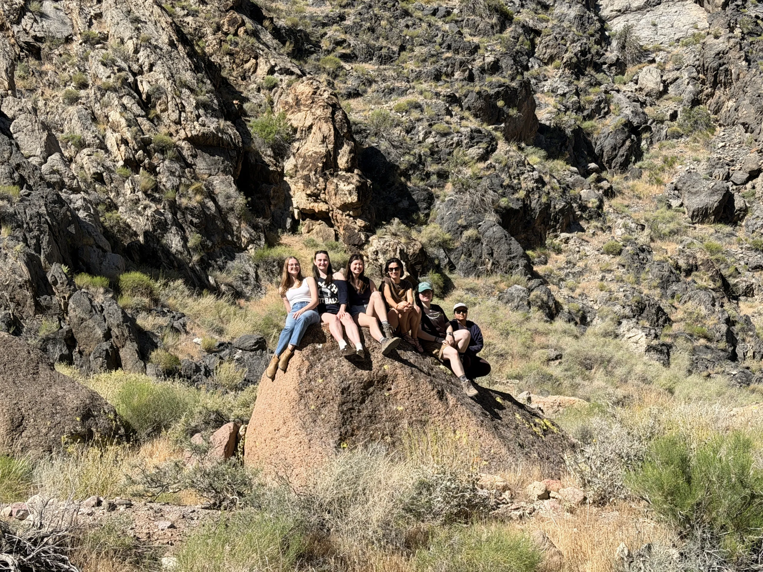 A group of six people sits together in a line atop a large brown boulder in a rugged, rocky canyon. Behind them, steep, dark rocky slopes with sparse green shrubs extend upward into the background.