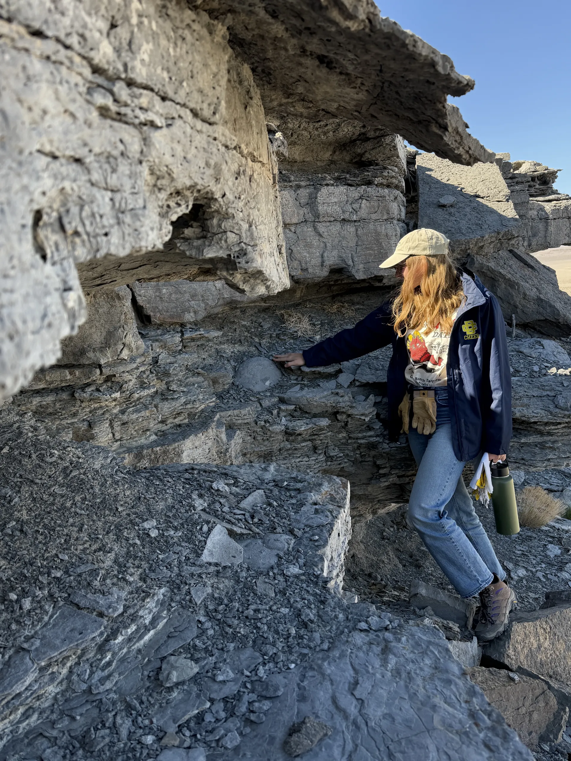 A person wearing a beige baseball cap, dark blue jacket, jeans, and hiking boots reaches out to touch a rounded rock feature embedded in a layered grey rock outcrop. She holds a green water bottle in her other hand beneath a clear blue sky.