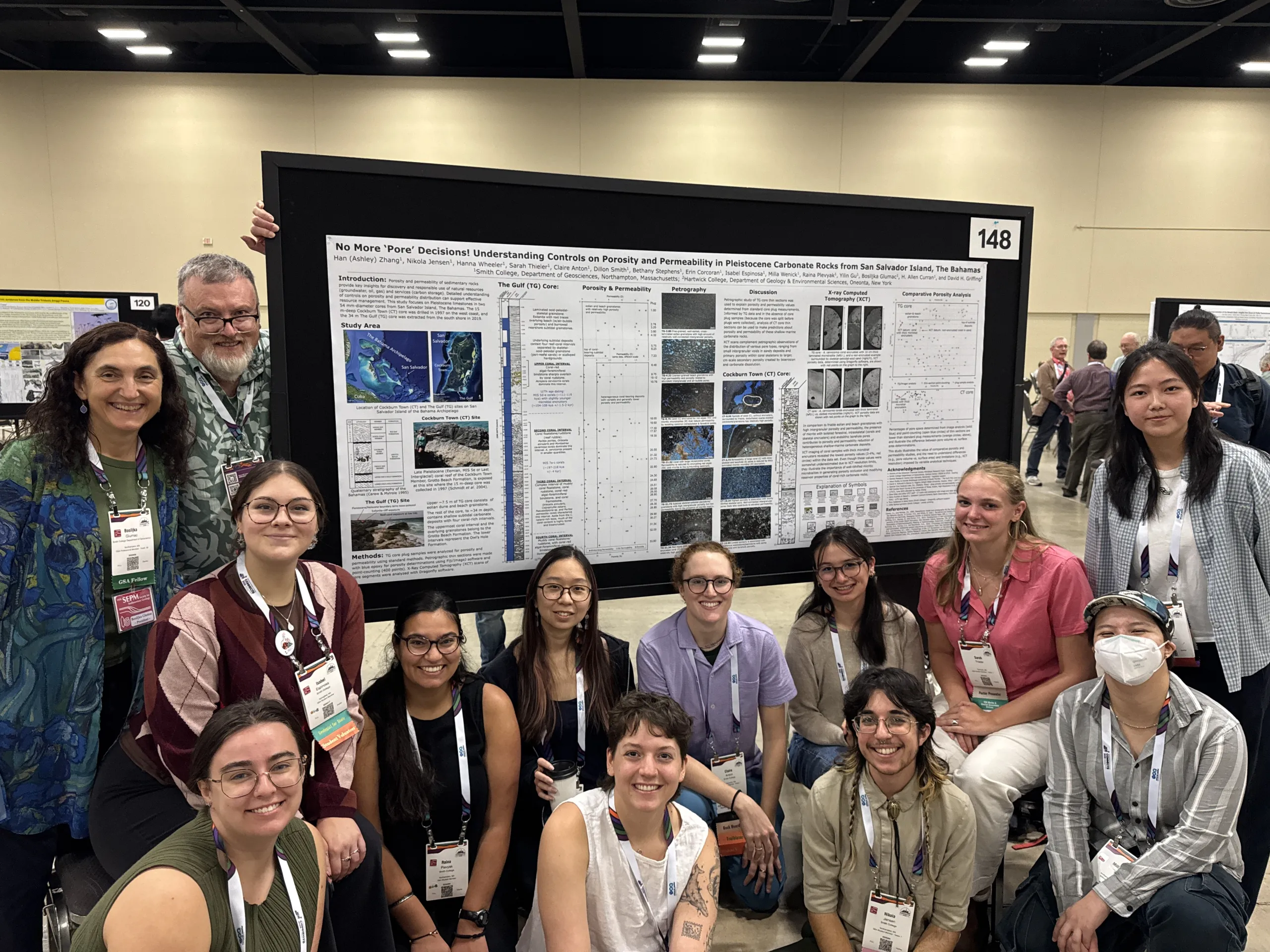 A large group of researchers and students pose together around a large research poster displayed on a black board numbered 148 at an academic conference. The poster is titled "No More 'Pore' Decisions! Understanding Controls on Porosity and Permeability in Pleistocene Carbonate Rocks from San Salvador Island, The Bahamas". Some people stand on either side of the display while others sit or kneel in the front row, many wearing name lanyards.