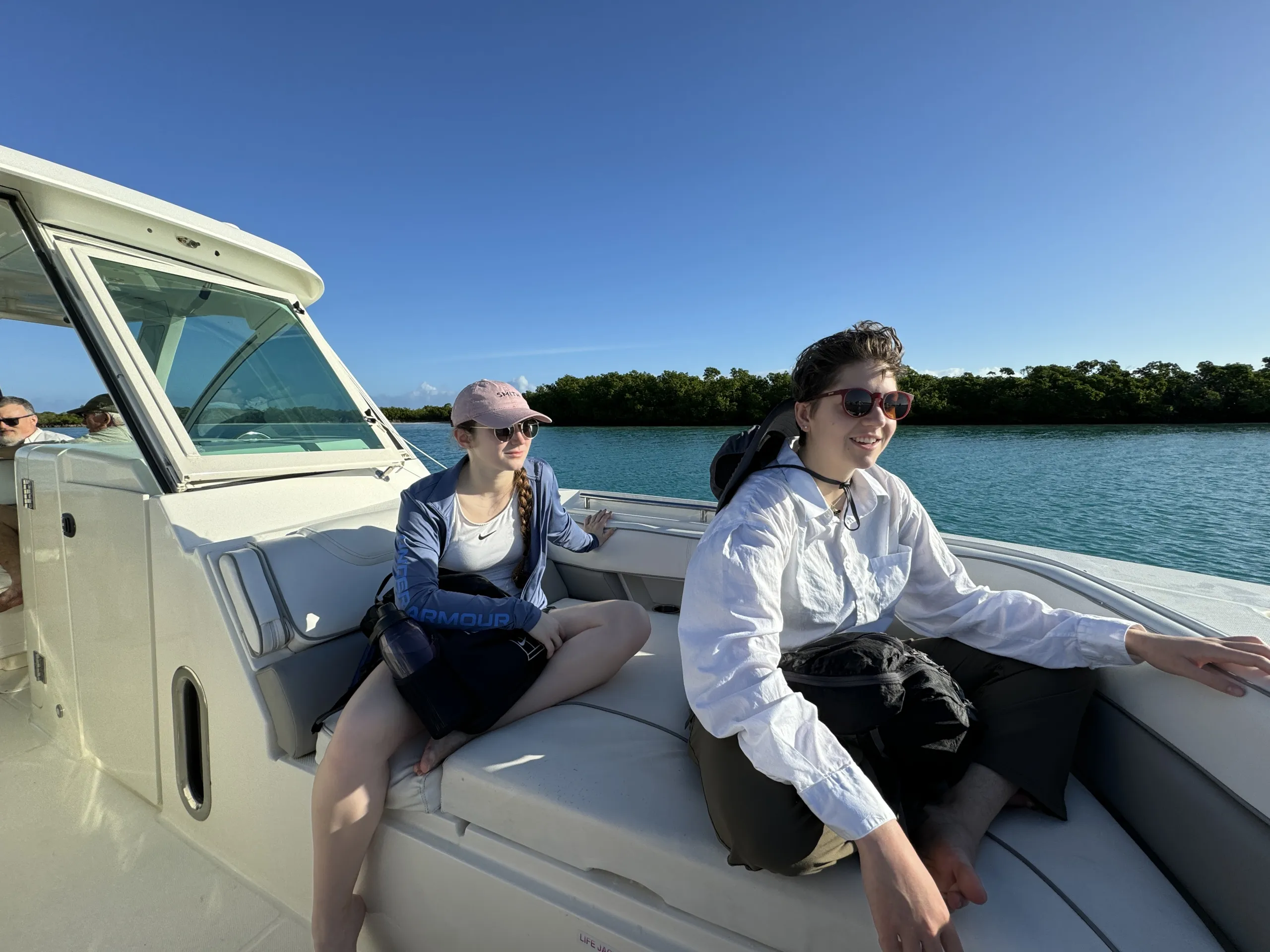 Two people sit comfortably cross-legged on the white cushioned bow seating of a boat cruising through calm, turquoise water. The person in the foreground wears sunglasses, a long-sleeved white button-down shirt, and dark pants. Behind them, the second person wears a pink baseball cap, sunglasses, and a long-sleeved blue jacket over a white top. Lush green foliage along the shoreline is visible under a clear, bright blue sky.