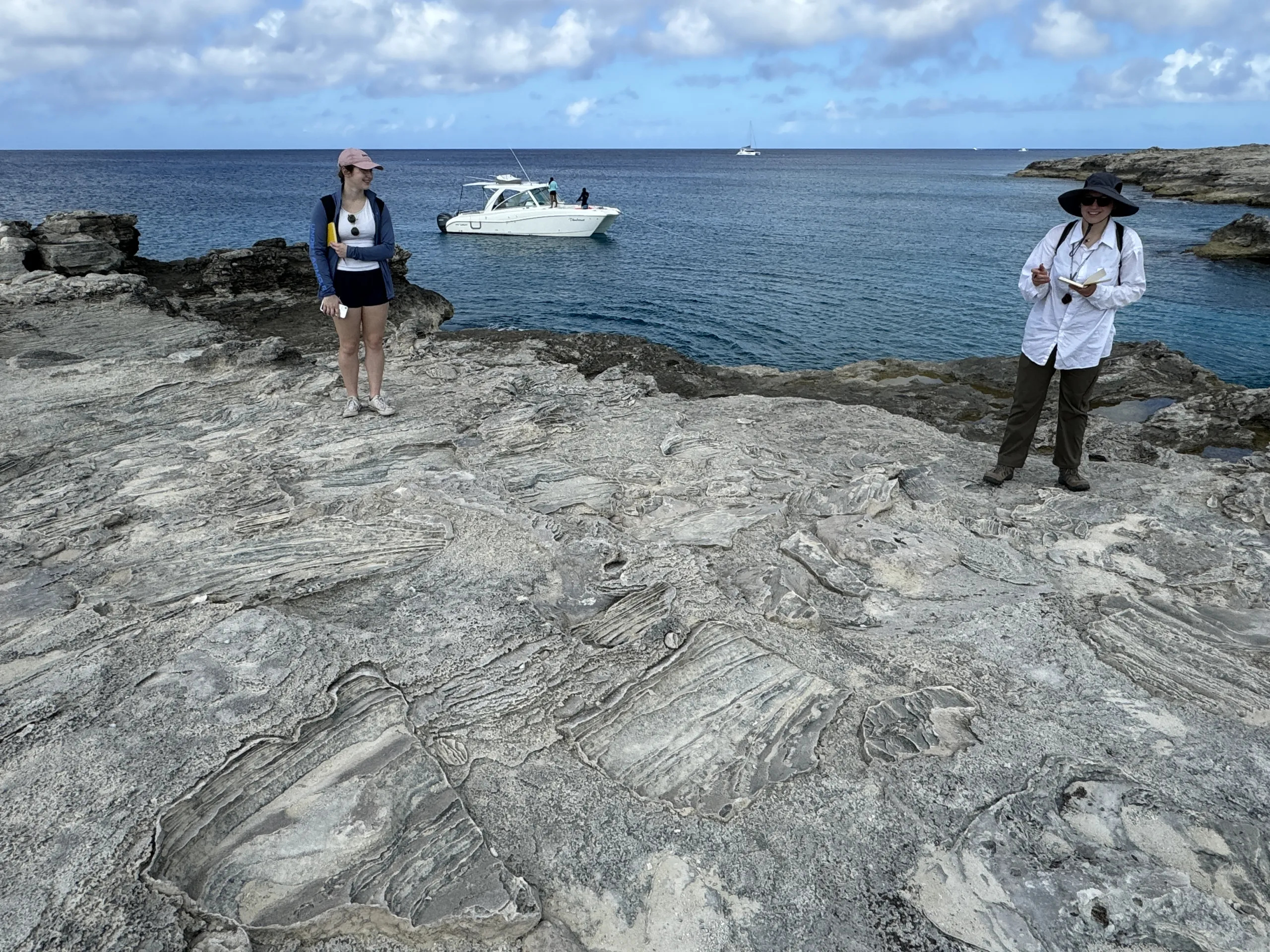 Two people stand on a broad, flat expanse of weathered, layered limestone coastal rock overlooking the sea. On the left, a person in a pink cap, blue jacket, and shorts stands holding a notepad. On the right, a person in a wide-brimmed sun hat, white shirt, and dark pants holds an open notebook and smiles toward the camera. A white motorboat is anchored offshore in deep blue water under a partly cloudy sky.
