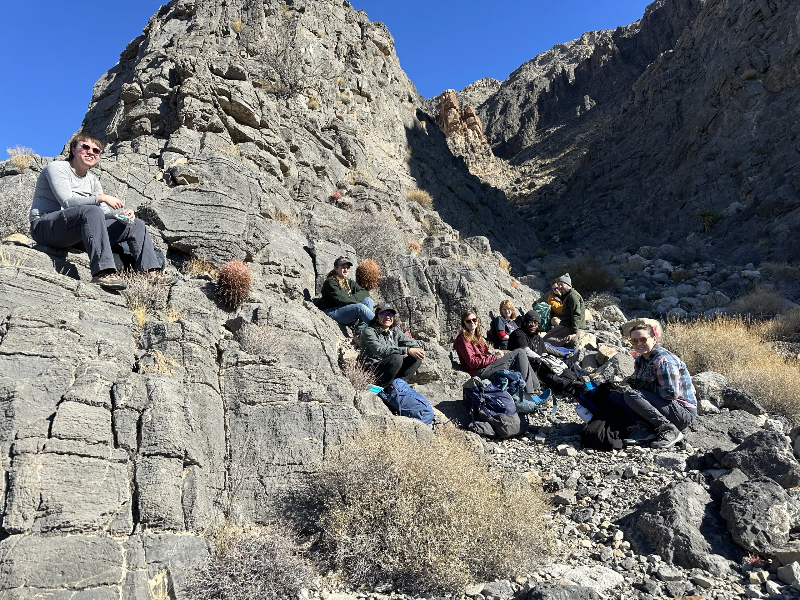 A group of hikers rests along a rocky, steep canyon path in a sunny desert landscape. Several people sit on large, layered grey rock outcrops on the left, while others sit on the gravelly canyon floor near backpacks on the right, with steep rock walls rising in the background under a clear blue sky.