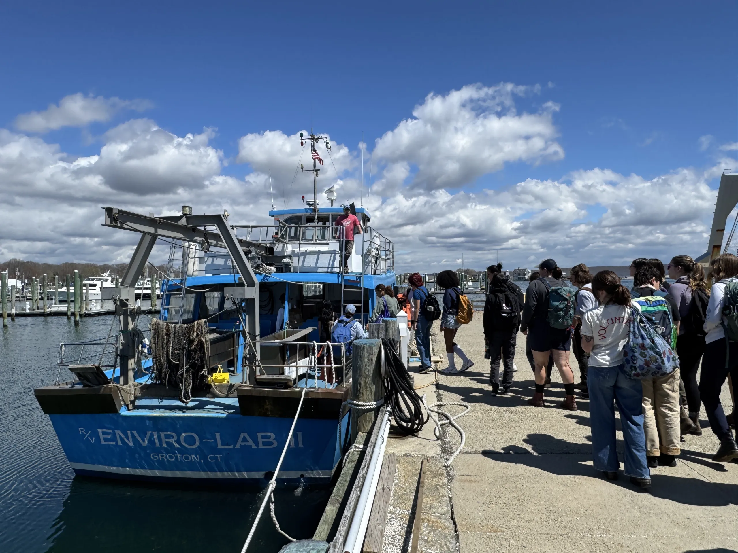 A group of people wearing backpacks stands along a wooden pier waiting to board a blue research vessel named "R/V ENVIRO-LAB II GROTON, CT". The boat features research equipment like a rear A-frame and rigging. The scene takes place under a bright blue sky filled with puffy white clouds, with calm water in the harbor.