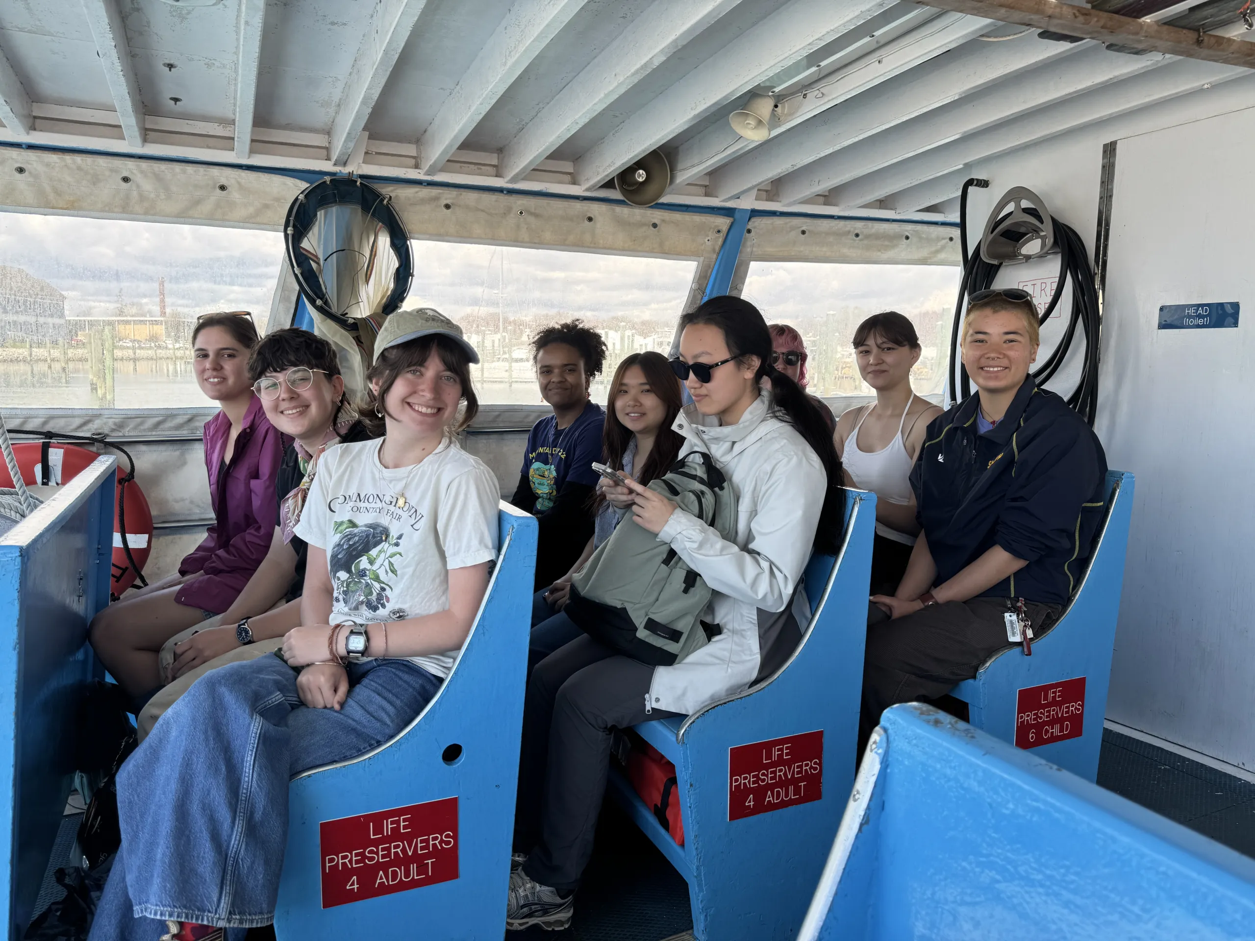 A group of students sits inside the covered passenger cabin of a boat with bright blue seats marked "LIFE PRESERVERS". They are smiling towards the camera, with large windows behind them showing a harbor view.