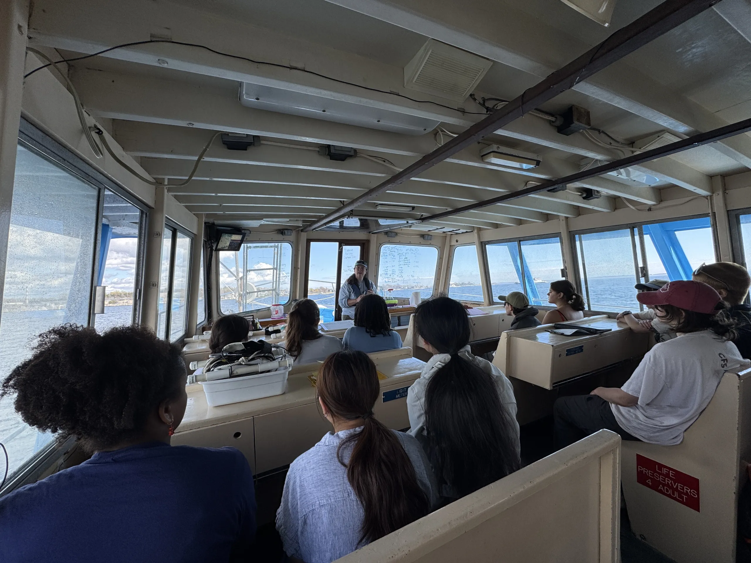 A group of students sits facing forward inside the cabin of a research vessel while an instructor stands near the bow speaking to the class. Large windows on all sides offer clear views of the surrounding open water under a sunny sky.