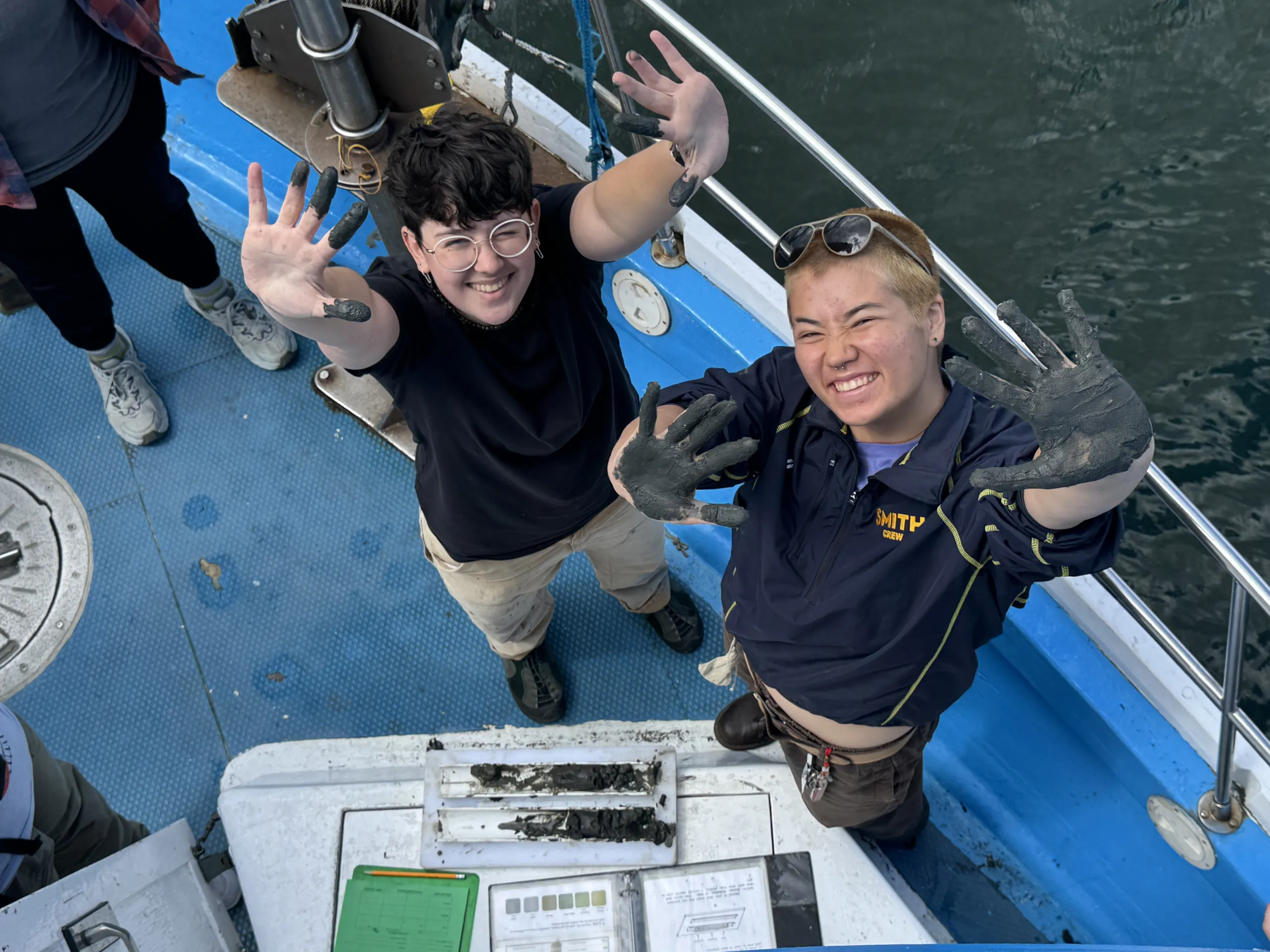 A high-angle view looking down at two students on the deck of a blue research boat, smiling and holding up both of their mud-covered hands toward the camera. Below them on a white table rests a tray containing dark sediment core samples next to a clipboard and record sheets.