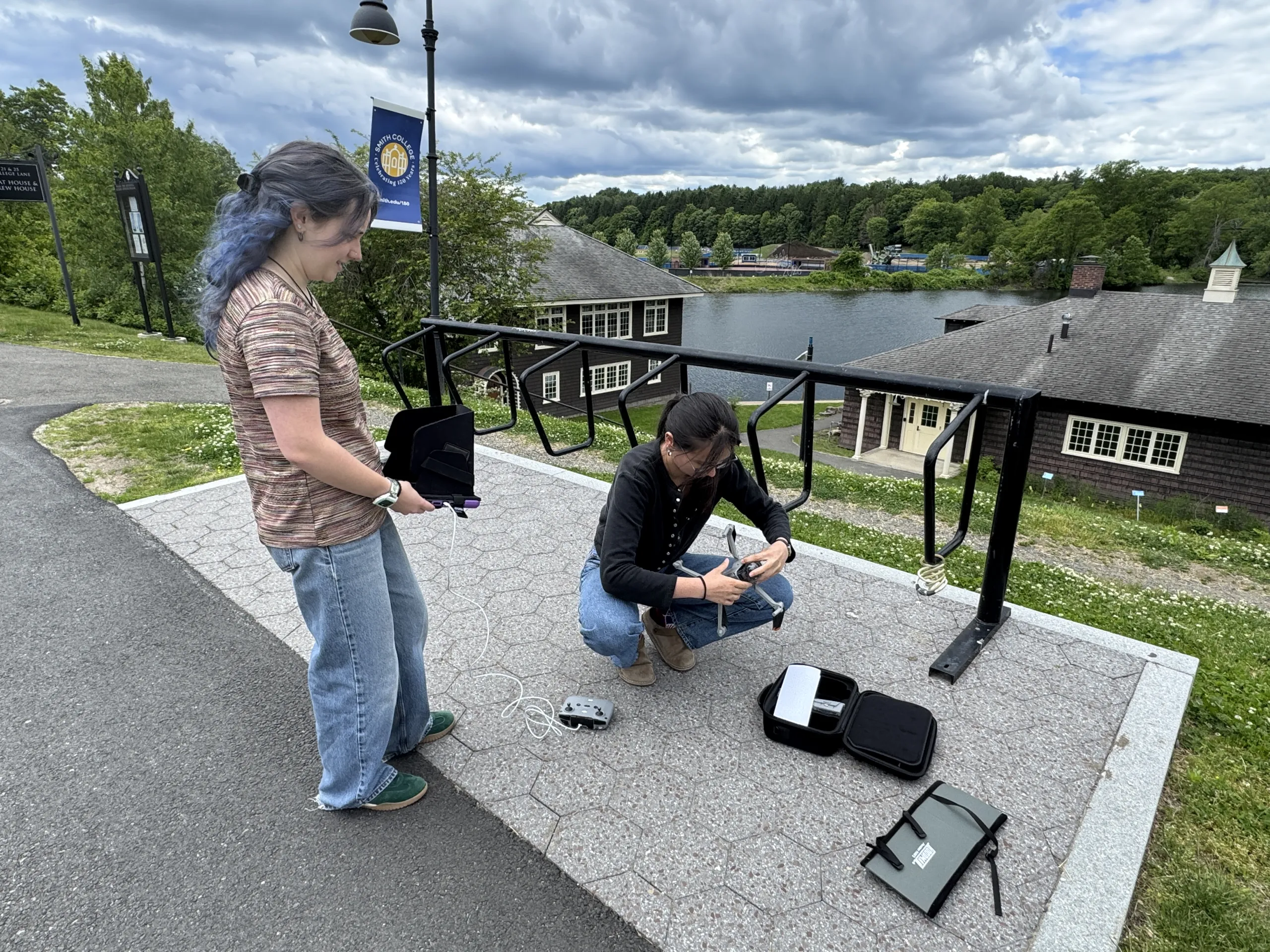 Two people work with a drone on a paved outdoor overlook above a lake. On the left, a person with blue-tinted hair stands holding a display screen attached to a remote controller. Next to them, a second person crouches down on the ground to inspect and adjust a small quadcopter drone beside open equipment cases. Campus buildings, trees, and water are visible in the background under a cloudy sky.