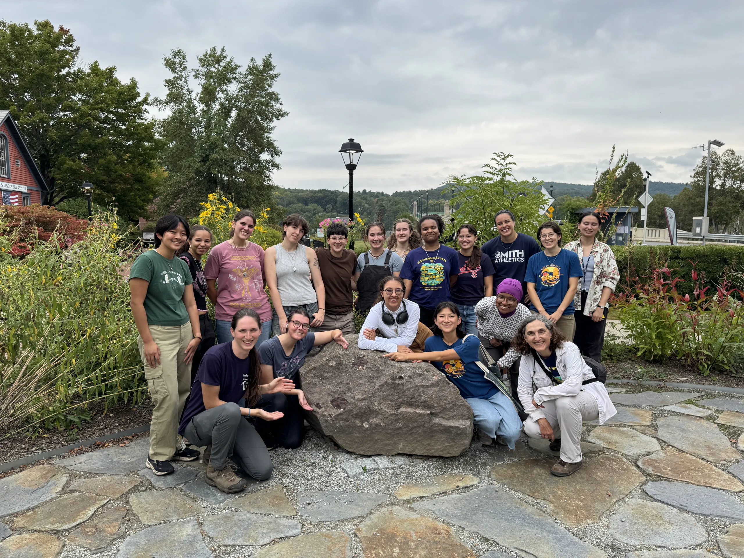 A large group of sixteen people gathers around a prominent brown rock set on a flagstone patio outdoors. Some members stand in a row behind the rock, while others crouch or sit near it, gesturing toward the stone. Lush green foliage, flowering plants, and trees surround the area under an overcast sky.