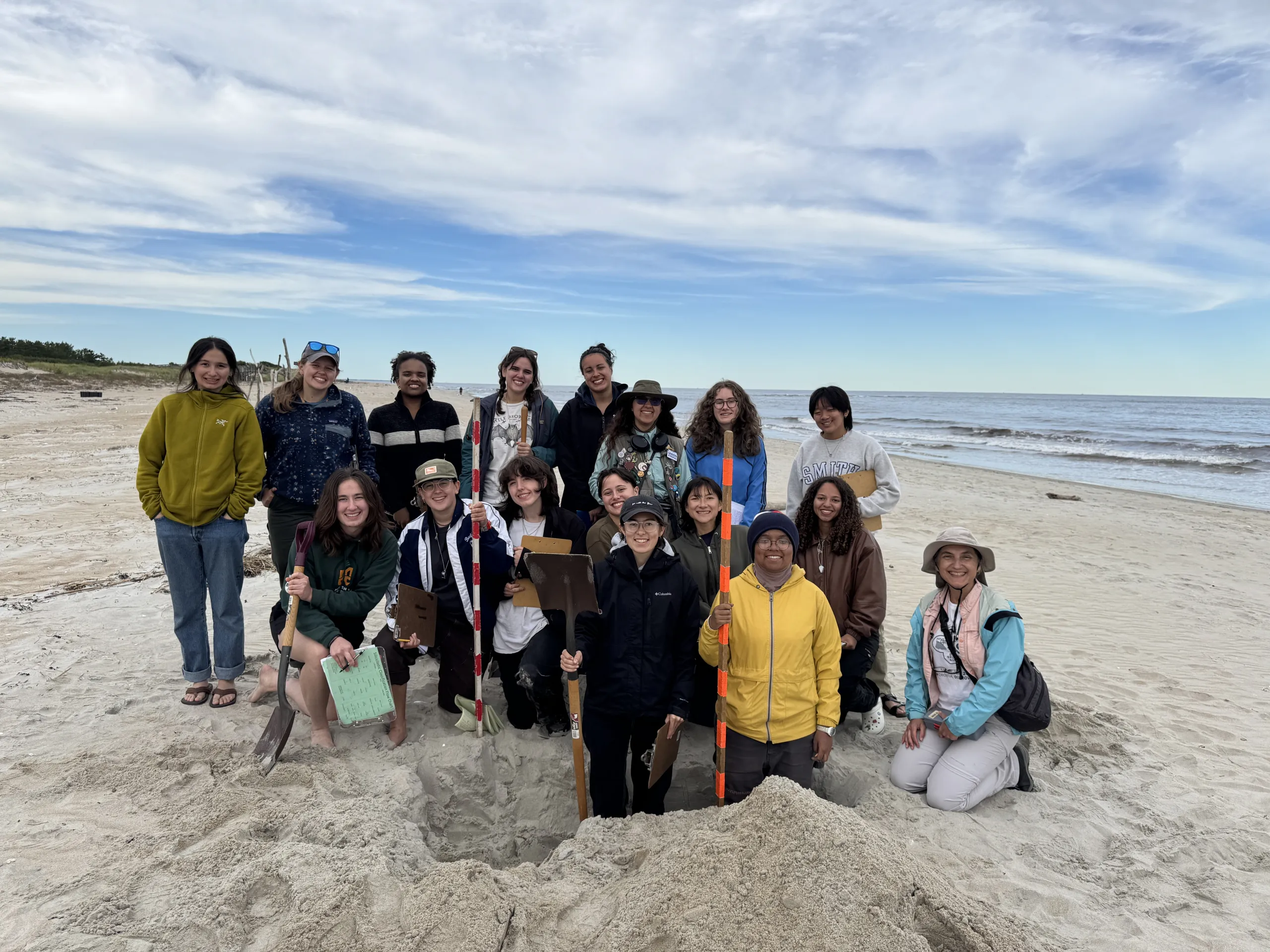 A group of researchers posing around a pit dug into a sandy beach, holding shovels, clipboards, and surveying poles with the ocean and a cloudy blue sky in the background.