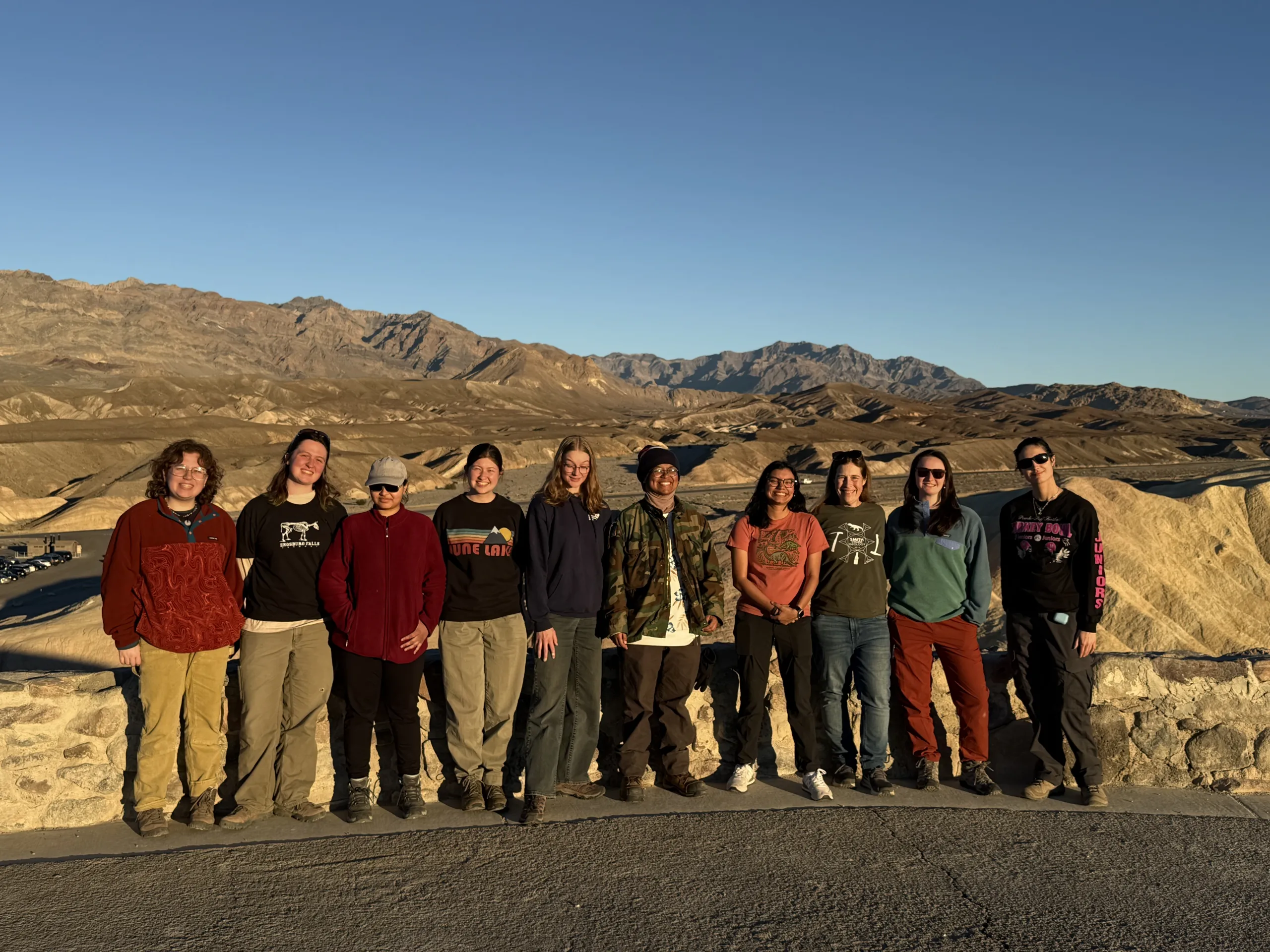 A group of ten people stands lined up along a low stone wall at an scenic overlook under a clear blue sky. Vast, sunlit desert badlands and rugged mountain ranges stretch across the background.