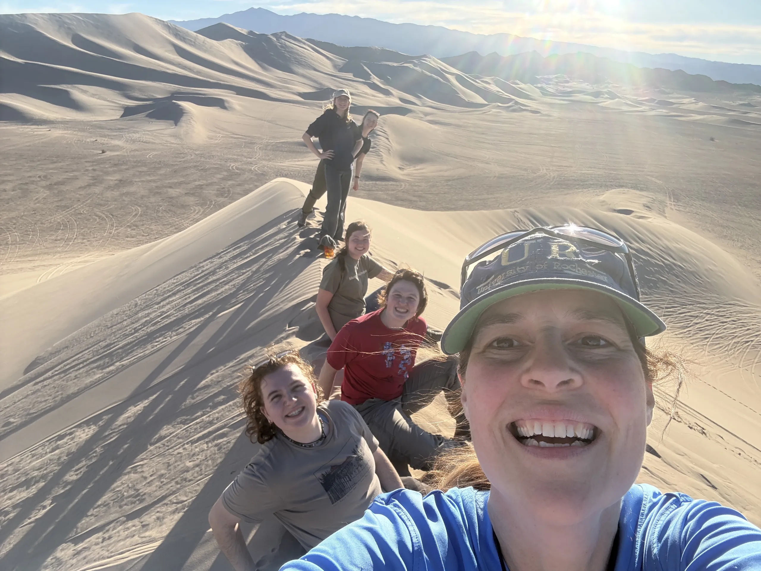 A high-angle group selfie taken along the narrow ridge of a large sand dune. Six people are positioned in a line trailing back along the crest of the dune, with wide sand fields and distant mountains visible under a bright, sunny sky.