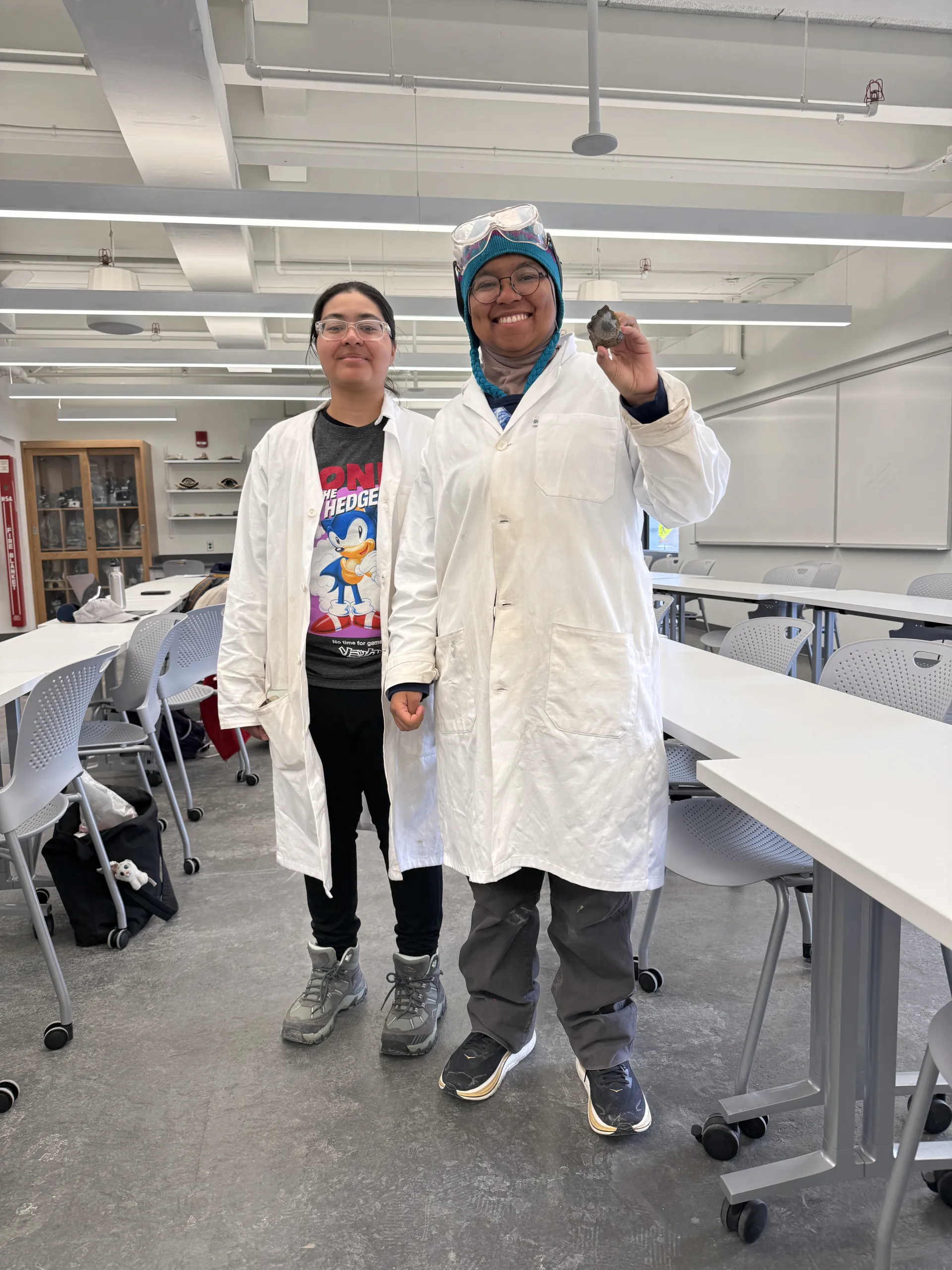 Two researchers in white lab coats stand inside a modern laboratory classroom with rows of white desks and chairs. The person on the right, wearing safety goggles over a colorful knit cap, smiles while holding up a small dark rock specimen for the camera. The person on the left stands beside them wearing clear protective glasses over a graphic shirt.