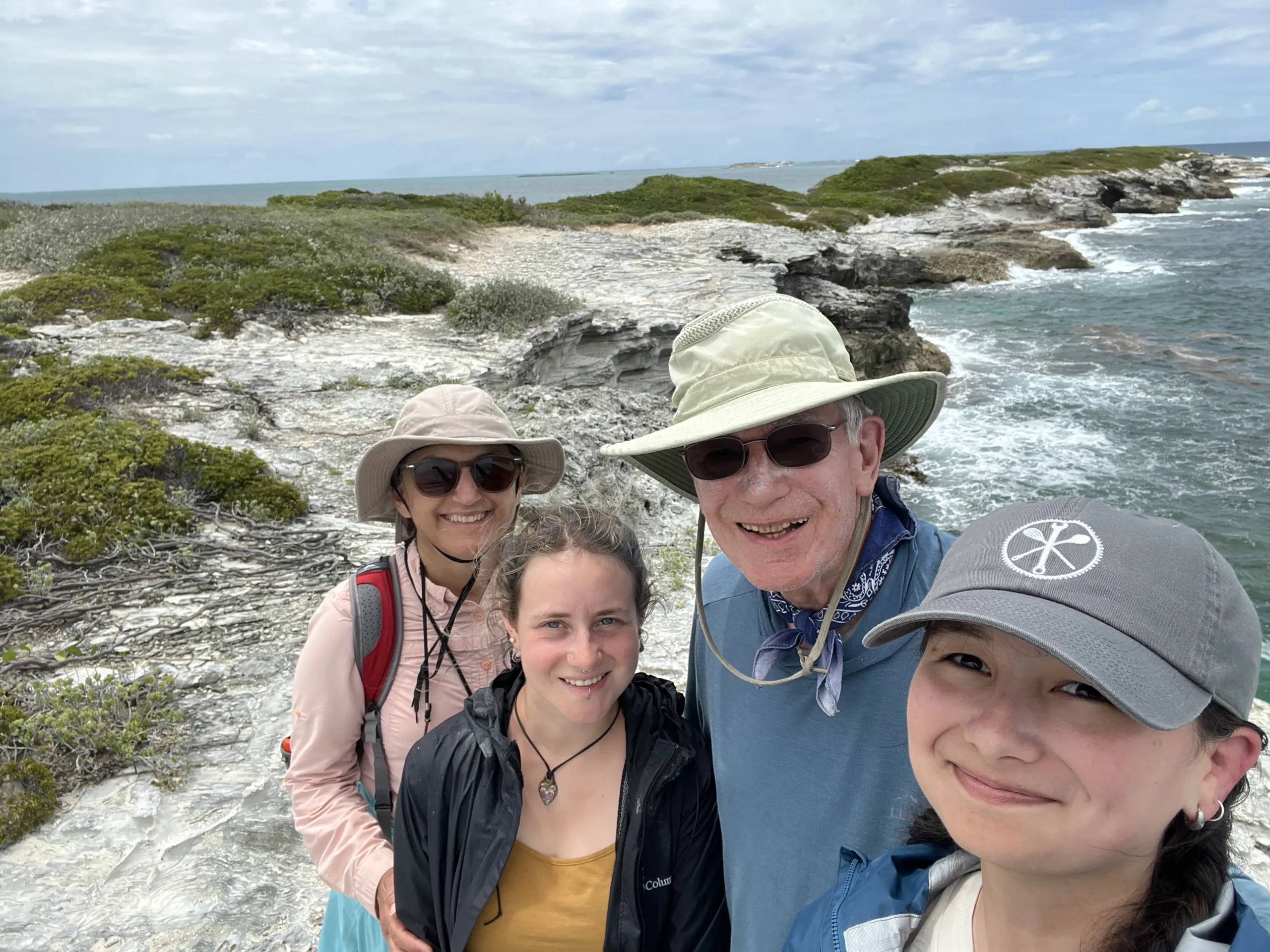 Four researchers smile for a close selfie on a rocky coastal cliff. The group includes a person on the far right holding the camera while wearing a grey baseball cap, a man in a wide-brimmed sun hat and sunglasses, a person in a black Columbia jacket, and a person in a tan sun hat and pink shirt. Behind them, rugged white limestone rock formations lead down to turquoise ocean water under an overcast sky.