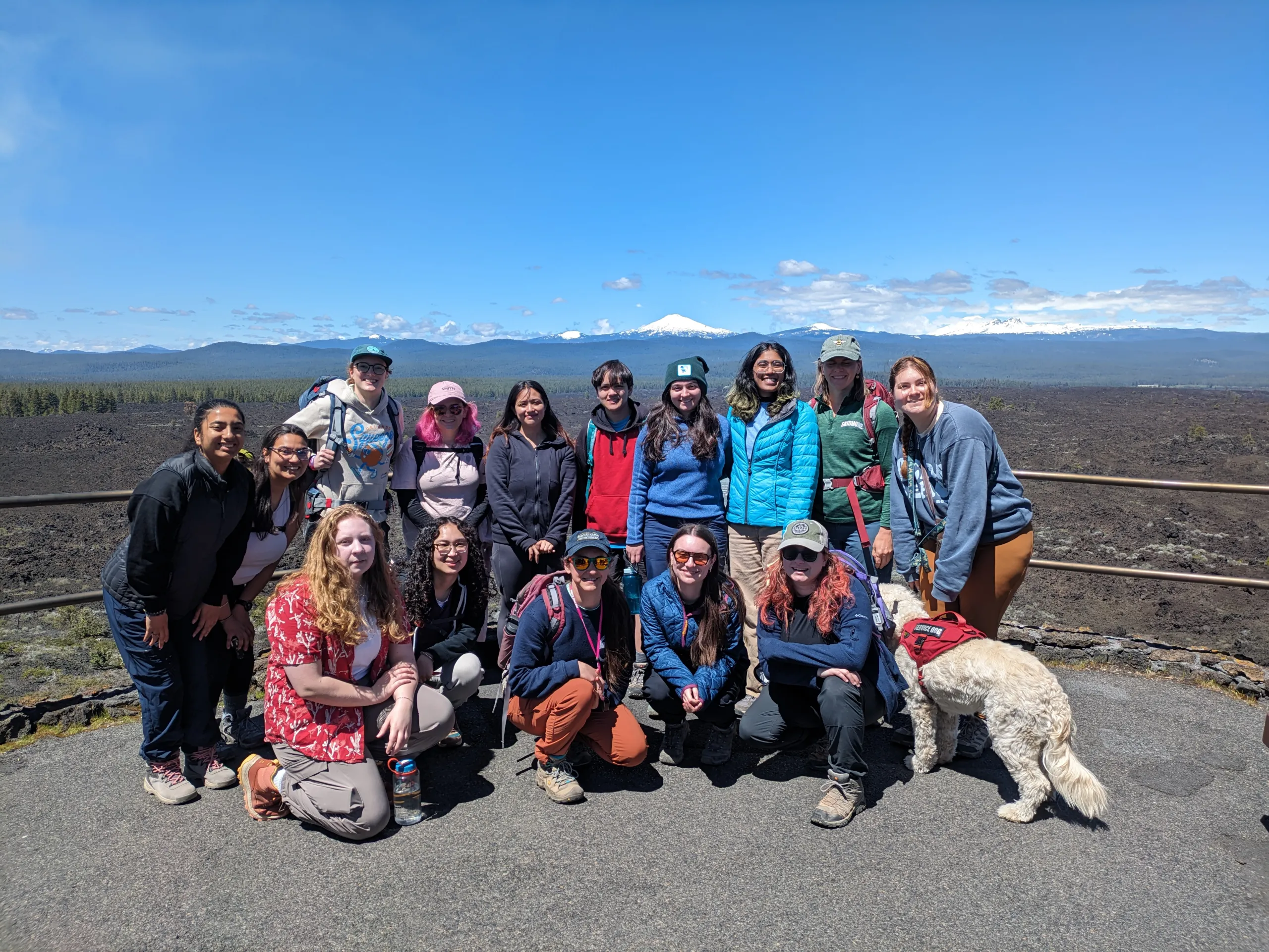 A group of fourteen people and a light-colored dog wearing a red vest pose together on a paved outlook under a bright blue sky. Some people are kneeling or crouching in the front row while others stand in the back. Behind them, a dark volcanic rock landscape stretches toward a distant line of mountains with snow-capped peaks.