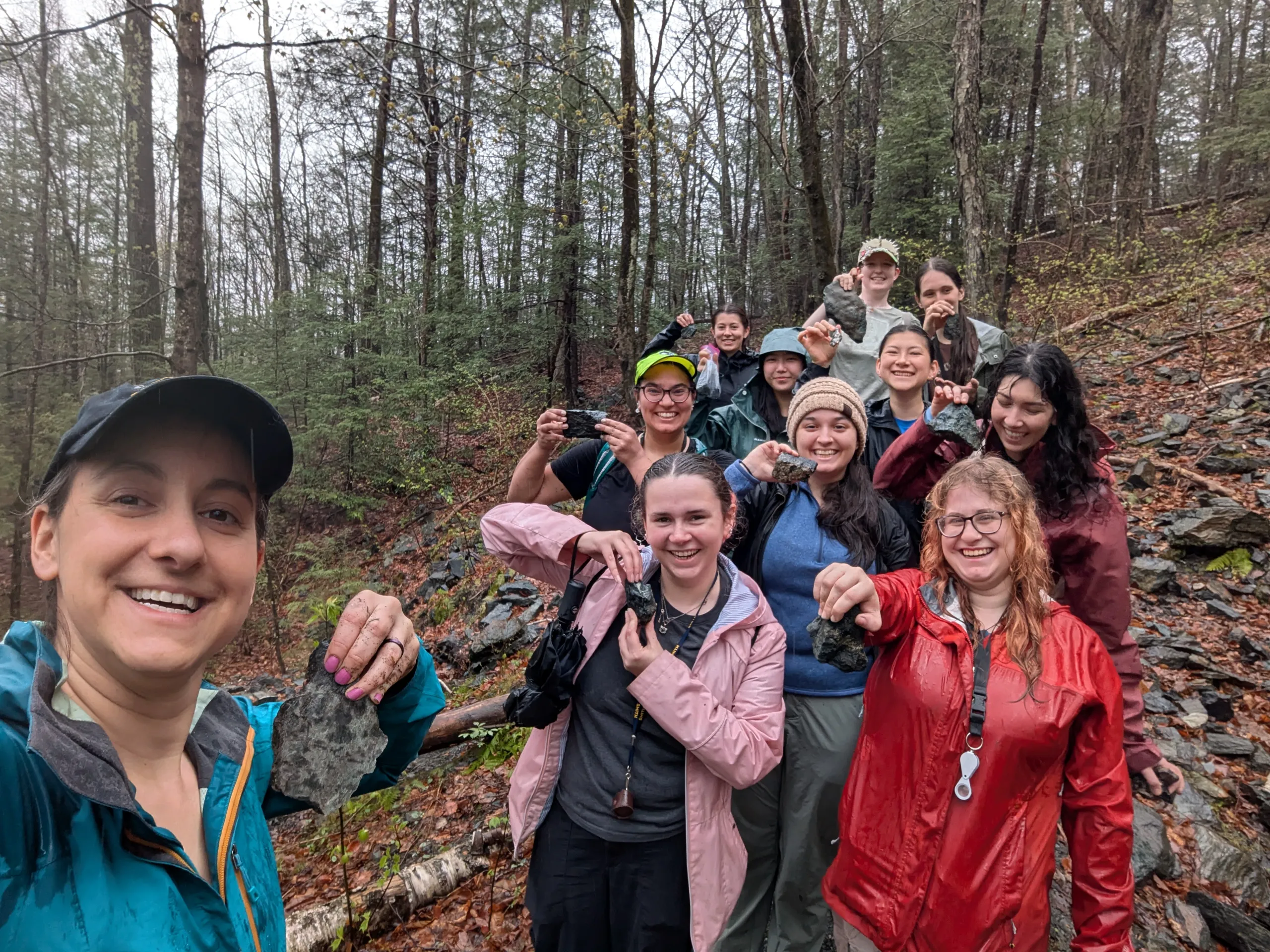 A group of ten people pose for a group selfie on a sloped, wooded trail in a forest. Many of them hold up dark mineral or rock specimens toward the camera. They are dressed in rain jackets and casual outdoor wear, surrounded by bare and early-spring trees.