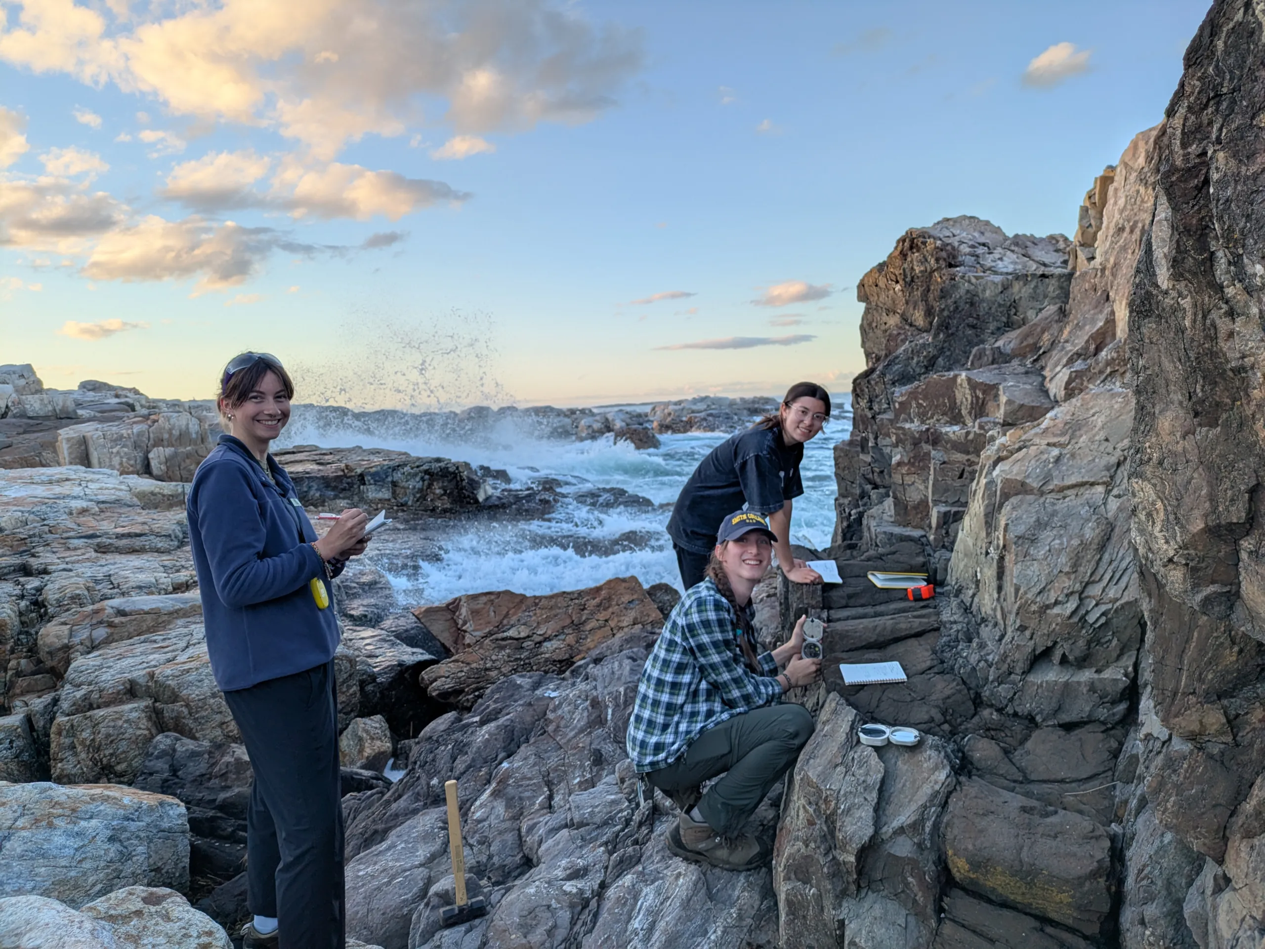 Three researchers conduct fieldwork on a rugged, rocky coastal formation as ocean waves crash against the rocks in the background. One person stands on the left holding a notepad, a second person kneels near a rock face using a geological compass and writing notes, and a third person stands near the rocks looking toward the camera. The sky above shows soft evening sunlight on scattered clouds.