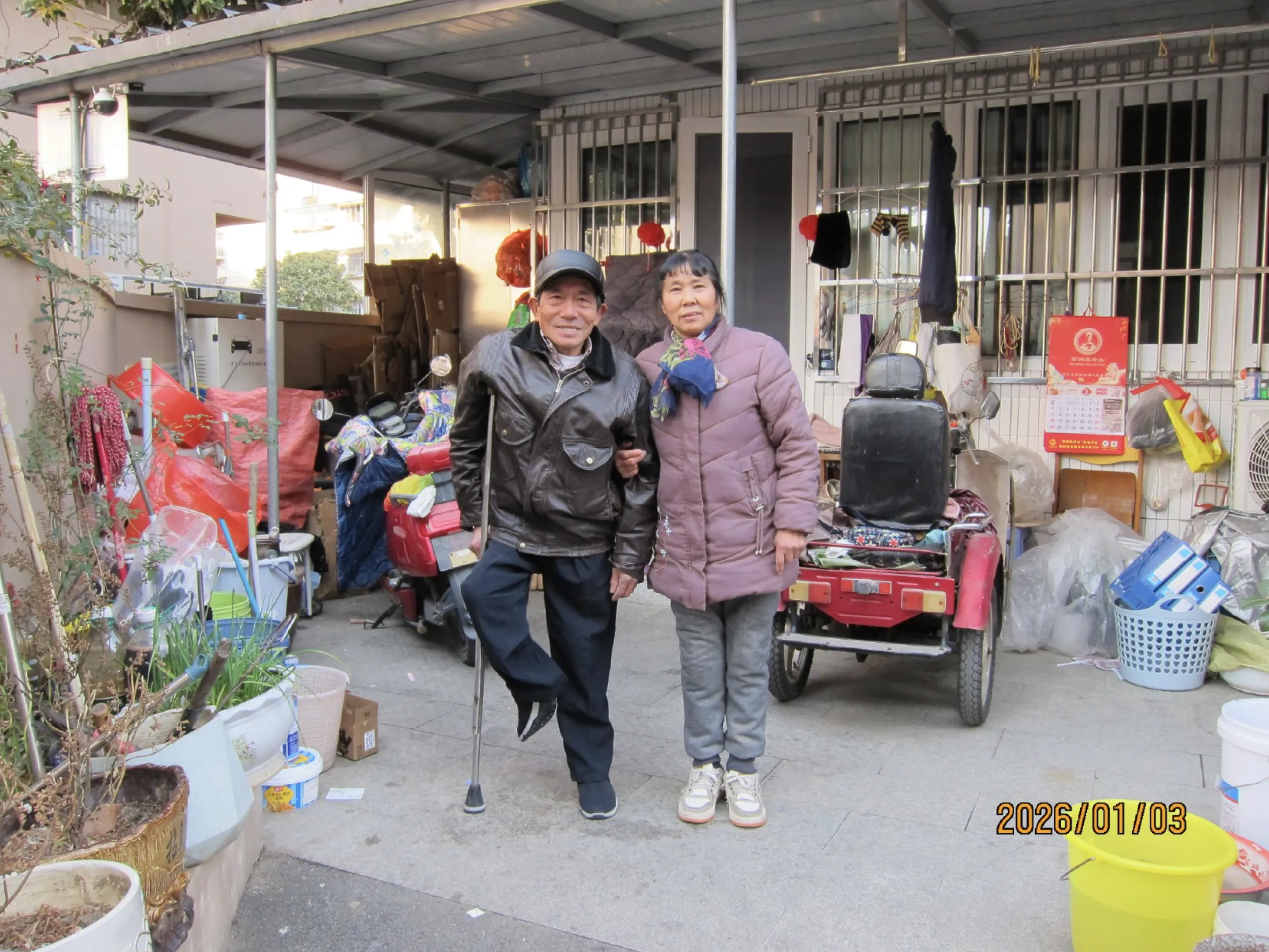 Two people in China lean on each other and smile at the camera. The man is on crutches.