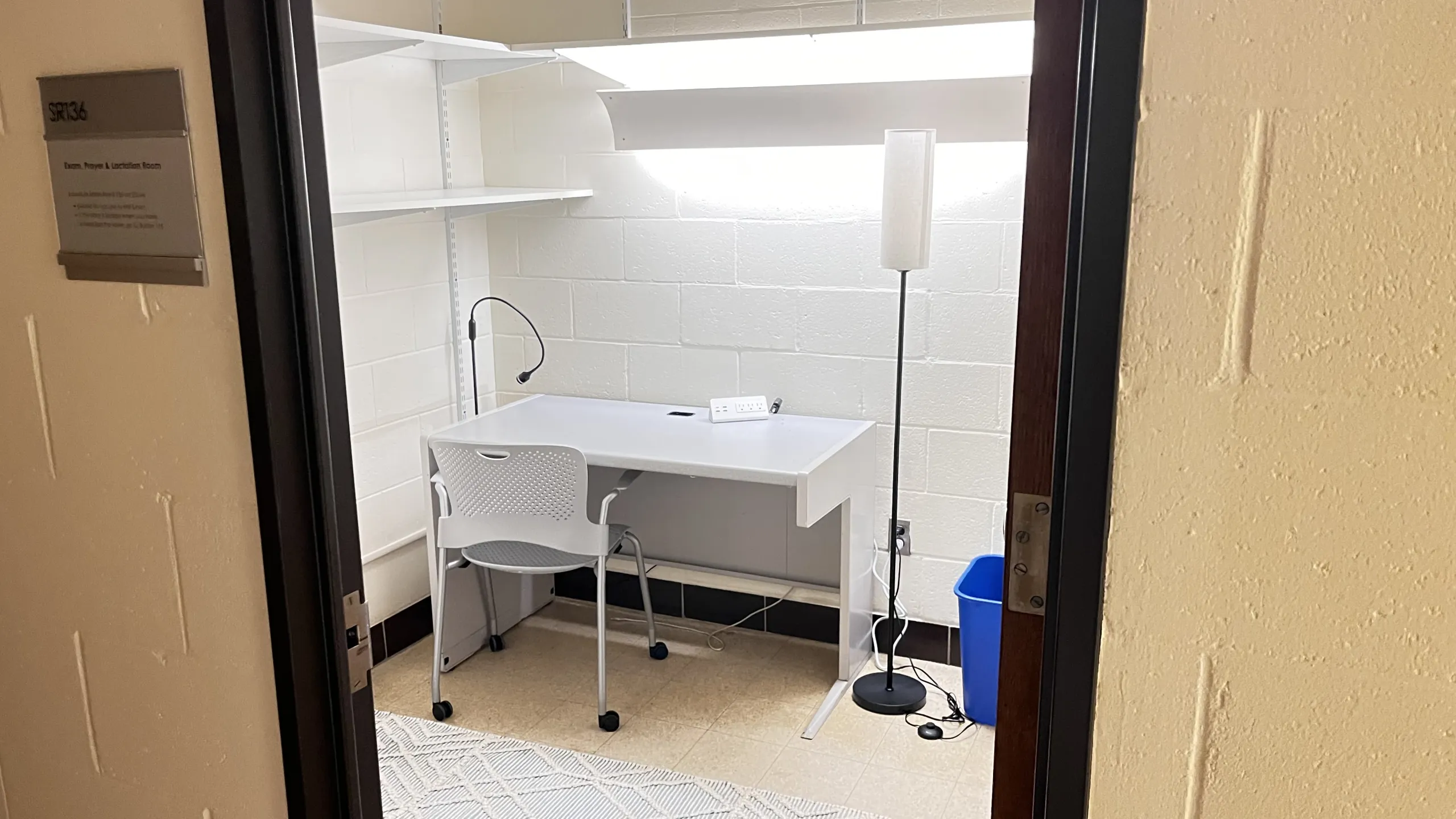 A small, well-lit lactation room with a white chair at a desk, shelves above, a floor lamp beside, a wall-mounted sign outside, and a blue bin on the floor.