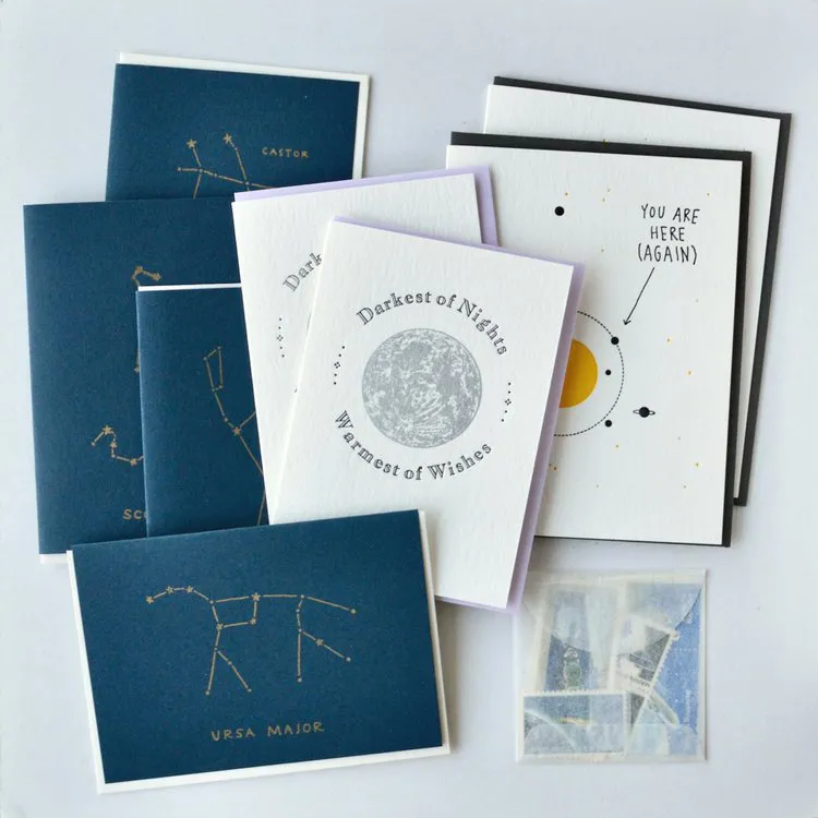 Five blue cards, four white cards, and a package of vintage stamps, which are found in the Space Assortment! greeting card bundle from Squeezebox Press by Smith College alum Emma Swartz
