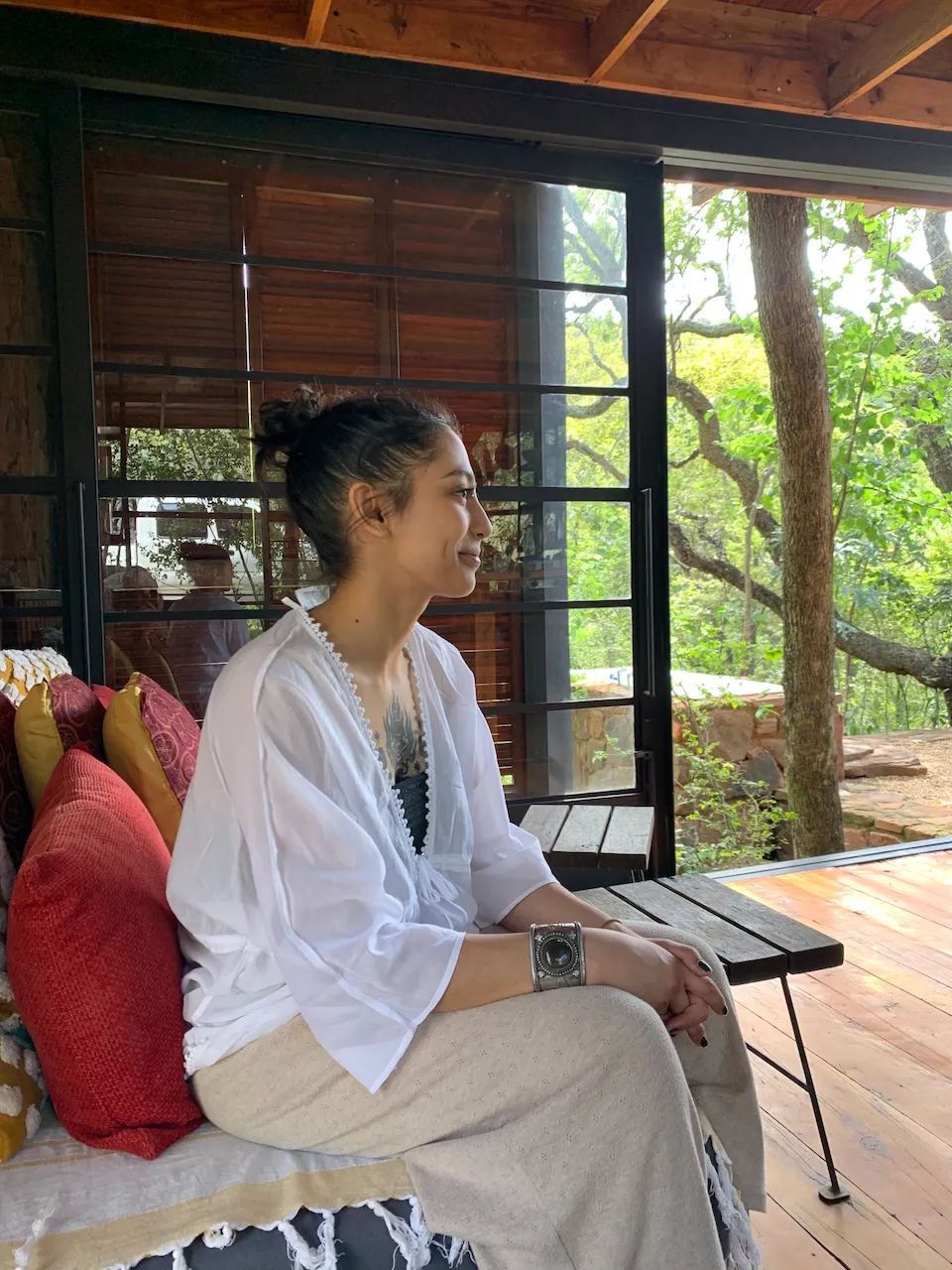 Tiki Pather sits on a chair in a wooden paneled room with a sliding door open to greenery