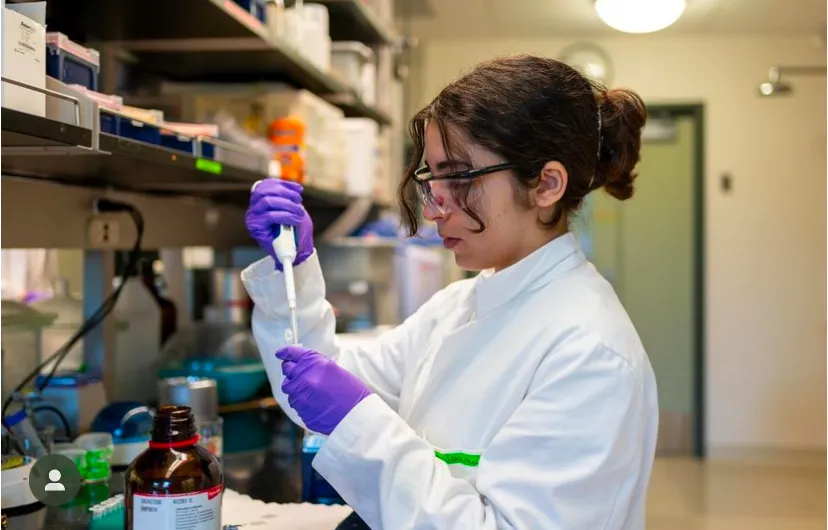 Karen Bekhazi in white lab coat and goggles using scientific instruments