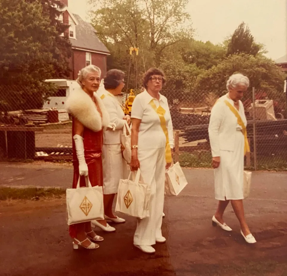Four Smithies in 1974 at Reunion, wearing white with yellow sashes. One alum is wearing a red dress with a white fur stole.