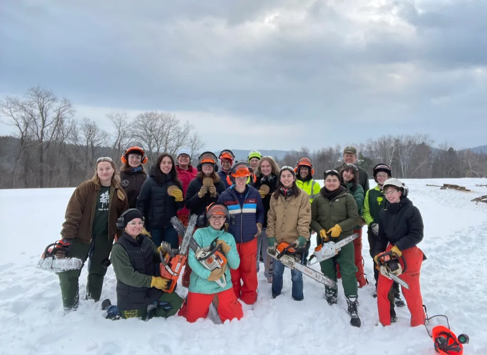 Workshop attendees pose for a group photo, wearing bright safety gear and some holding chain saws in the snow.