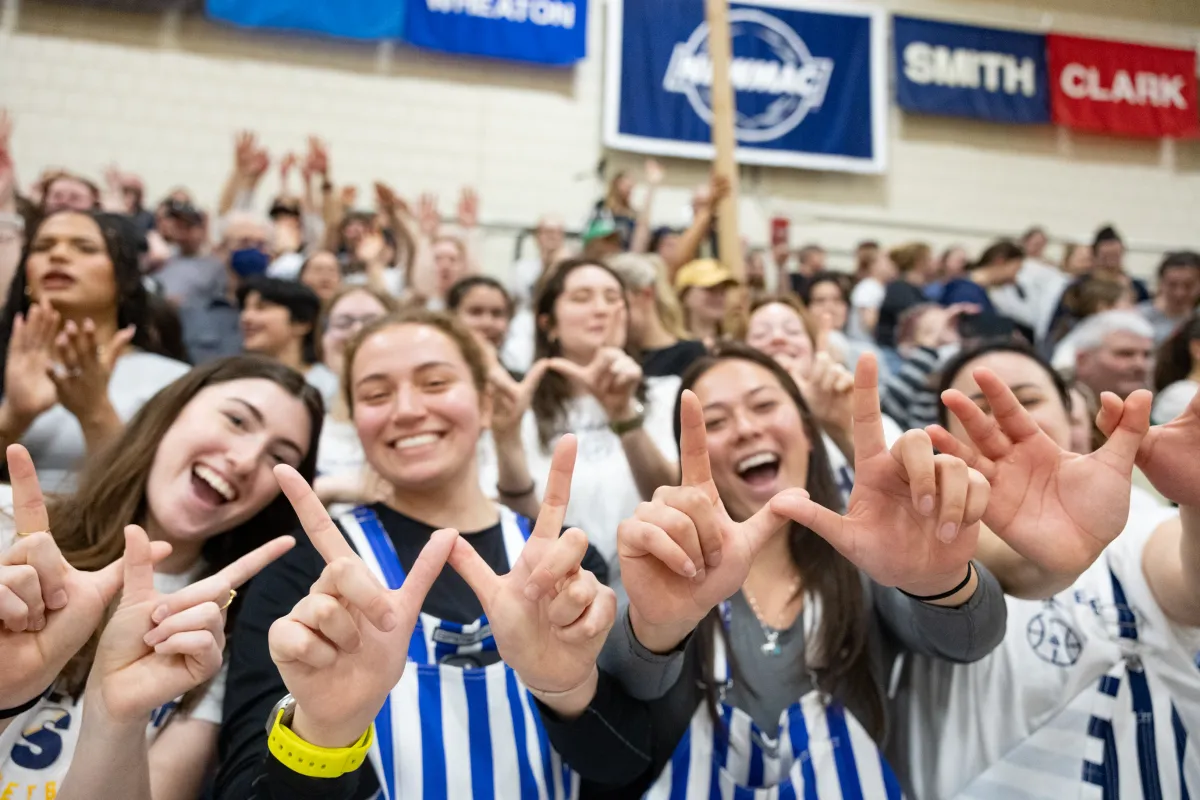 Students cheer excitedly in crowded gymnasium bleachers, making hand signs toward the camera.