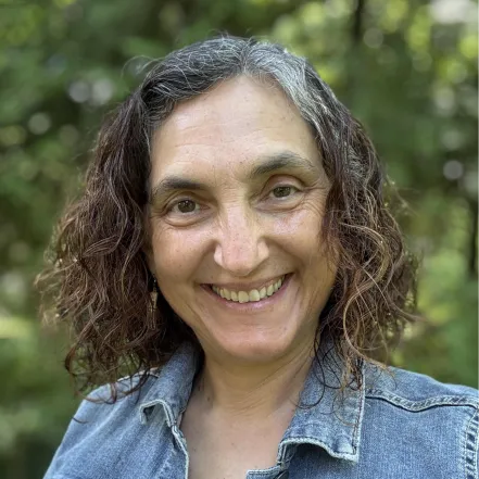 A person with curly brown and gray hair is smiling at the camera, wearing a denim jacket, with green foliage blurred in the background.